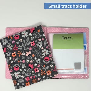 May include: A small, pink tract holder with a floral patterned cover. The cover features a dark gray background with pink, orange, and white flowers and paw prints. The holder contains a green and white tract with the word "Tract". The text "Small tract holder" is in the top right corner.