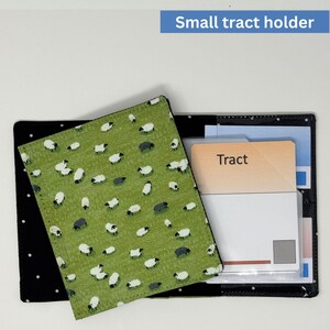 May include: A small tract holder with a green cover featuring a sheep pattern. The holder is open, revealing a card labeled "Tract" and other inserts. The holder is black with white polka dots.