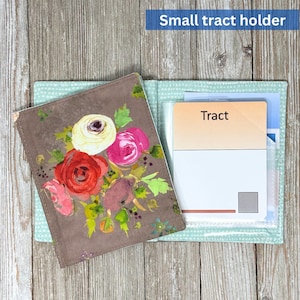 May include: A small tract holder with a floral design. The cover is brown with a watercolor painting of red, pink, and white roses. The interior is light blue with a polka dot pattern. The word "Tract" is visible on a card inside.