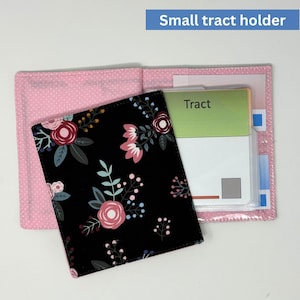 May include: A small tract holder, open to display its contents. The holder is pink with white polka dots and a black floral patterned cover. The text "Tract" is visible on a card inside the holder.
