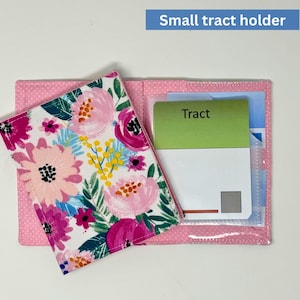 May include: A pink small tract holder with a floral design. The holder is open, revealing a clear pocket with a green card that says "Tract". The floral pattern includes pink, purple, and yellow flowers on a white background.