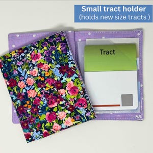 May include: A small, open tract holder with a floral print cover. The cover features a dark blue background with pink, red, and purple flowers. The interior is light purple with white polka dots. The holder contains a green and white tract.