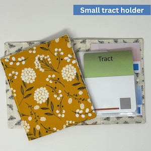 May include: A small tract holder with a mustard yellow floral cover and a light grey bee-patterned interior. The holder is open, revealing a card with the word "Tract" and other papers.