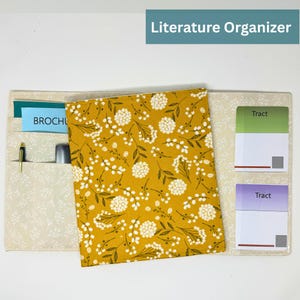 May include: A fabric literature organizer with a mustard yellow floral pattern cover. The organizer has multiple pockets, including one with a brochure and pen. The text "Literature Organizer" is displayed at the top.