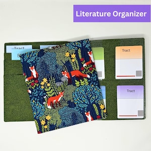 Può includere: Un organizer per la letteratura verde con una copertina blu navy raffigurante una scena di foresta con volpi arancioni, fiori gialli e fogliame verde. L'organizer ha più tasche e il testo "Literature Organizer".