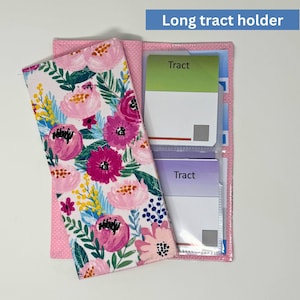 May include: A floral long tract holder with a pink polka dot backing. The front cover features a white background with pink and fuchsia flowers and green leaves. The holder is open, revealing cards with the word "Tract" on them.