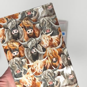 May include: A fabric item featuring a repeating pattern of Highland cows in various shades of brown, gray, and white. The cows have long, shaggy fur, horns, and pink noses. The item is held in a hand, with a white background.