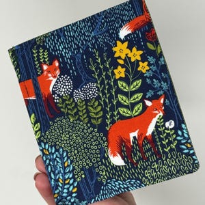 Puede incluir: Un artículo de tela azul marino con un estampado repetido de zorros naranjas, flores amarillas y follaje verde. El diseño incluye acentos blancos, azules y verdes, creando una escena forestal caprichosa. El artículo se sostiene en una mano.