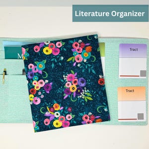 Puede incluir: Un organizador de literatura de tela color turquesa con un estampado floral. El organizador tiene bolsillos para documentos y un bolígrafo. La cubierta principal presenta un fondo azul oscuro con flores coloridas. El texto "Literature Organizer" es visible.
