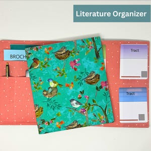 May include: A coral-colored literature organizer with white polka dots, featuring pockets for brochures, pens, and notepads. The central cover is teal with a bird and floral pattern. The text "Literature Organizer" is at the top.