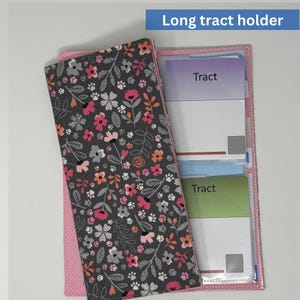 May include: A long tract holder with a floral pattern cover. The cover is dark gray with pink, orange, and white flowers. The holder is open, revealing three clear pockets with the word "Tract" on the cards.