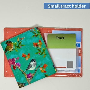 May include: A small tract holder with a teal and floral patterned cover featuring birds and flowers. The holder is open, revealing a clear pocket with a green card that says "Tract". The holder is orange with white polka dots.