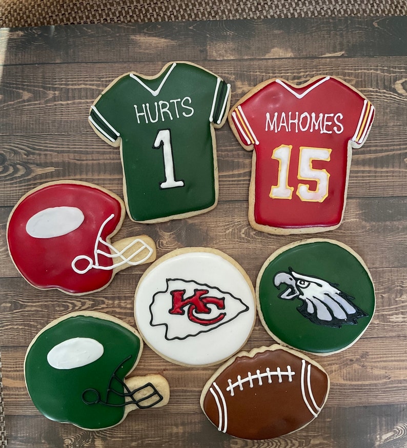 May include: Seven decorated sugar cookies, including two football jerseys, one with the name 'HURTS' and the number '1' in white on a green background and the other with the name 'MAHOMES' and the number '15' in white on a red background, two football helmets, one green and one red, a football, a white cookie with a red 'KC' logo and a green cookie with a white eagle.