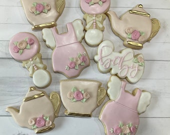 Puede incluir: Una colección de galletas decoradas con formas de peleles de bebé, teteras, tazas de té y sonajeros. Las galletas están glaseadas en tonos rosa pálido, crema y blanco, con detalles dorados y florales. Una galleta presenta la palabra baby.