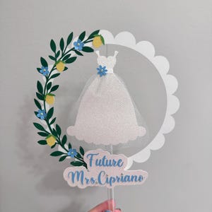 May include: White cake topper with a white dress silhouette, a green leafy garland with yellow lemons and blue flowers, and the text "Future Mrs. Cipriano".