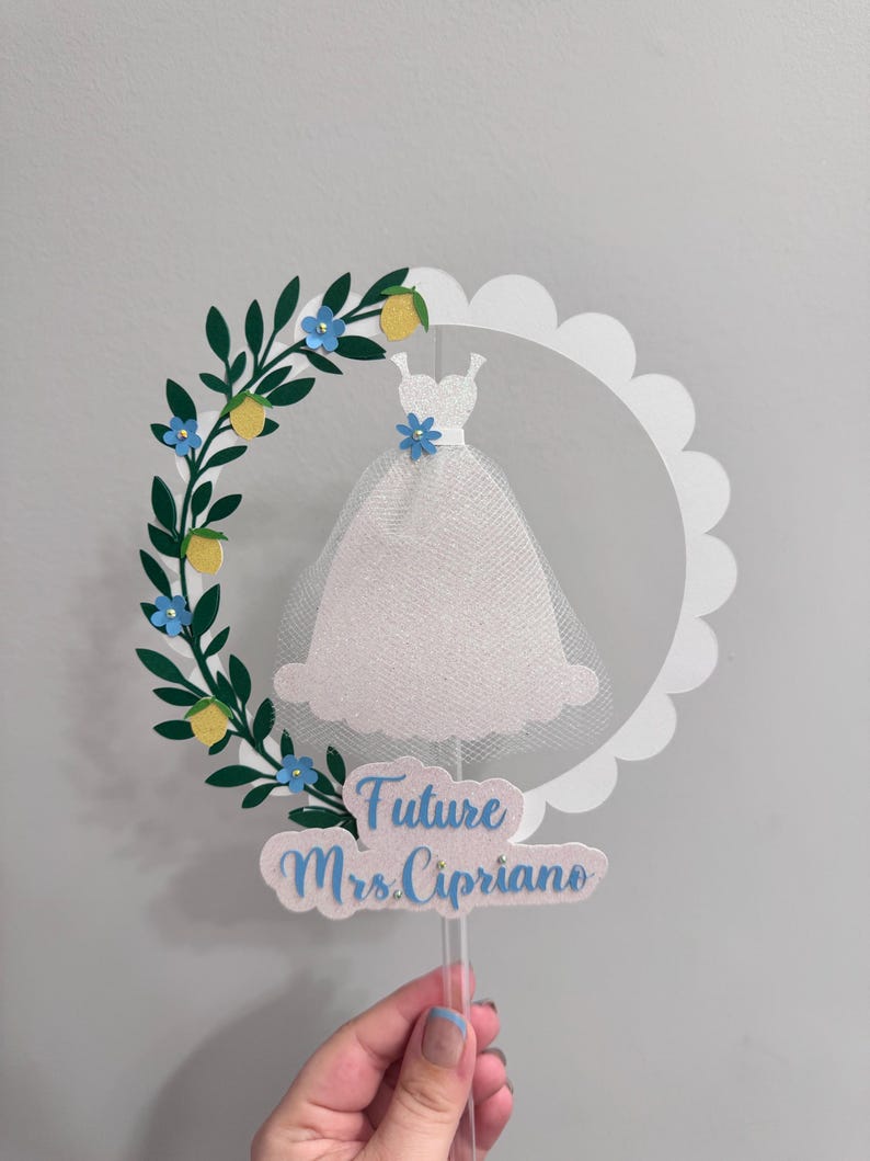 May include: White cake topper with a white dress silhouette, a green leafy garland with yellow lemons and blue flowers, and the text "Future Mrs. Cipriano".