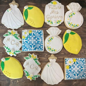 May include: A set of 12 decorated sugar cookies featuring a lemon theme. The cookies are decorated with white icing, yellow icing, green icing, blue icing, and gold accents. The cookies feature designs such as lemons, a wedding dress, a ring, a tile pattern, and the text "Future Mrs. Cipriano" and "MCR".
