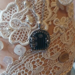 Pu&ograve; includere: Una collana con catena d'argento con un ciondolo a forma di porta verde. La porta &egrave; decorata con una cornice d'argento e una piccola manopola d'argento. Il ciondolo &egrave; attaccato a una catena d'argento.