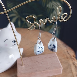 May include: A pair of white porcelain earrings with blue floral designs, hanging from a gold wire earring stand with the word "Smile" written on it.