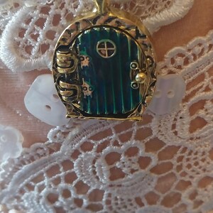 Pu&ograve; includere: Una collana con catena dorata con un ciondolo a forma di porta verde e dorata. La porta ha una piccola finestra rotonda e una maniglia dorata. La collana &egrave; su uno sfondo di pizzo bianco.