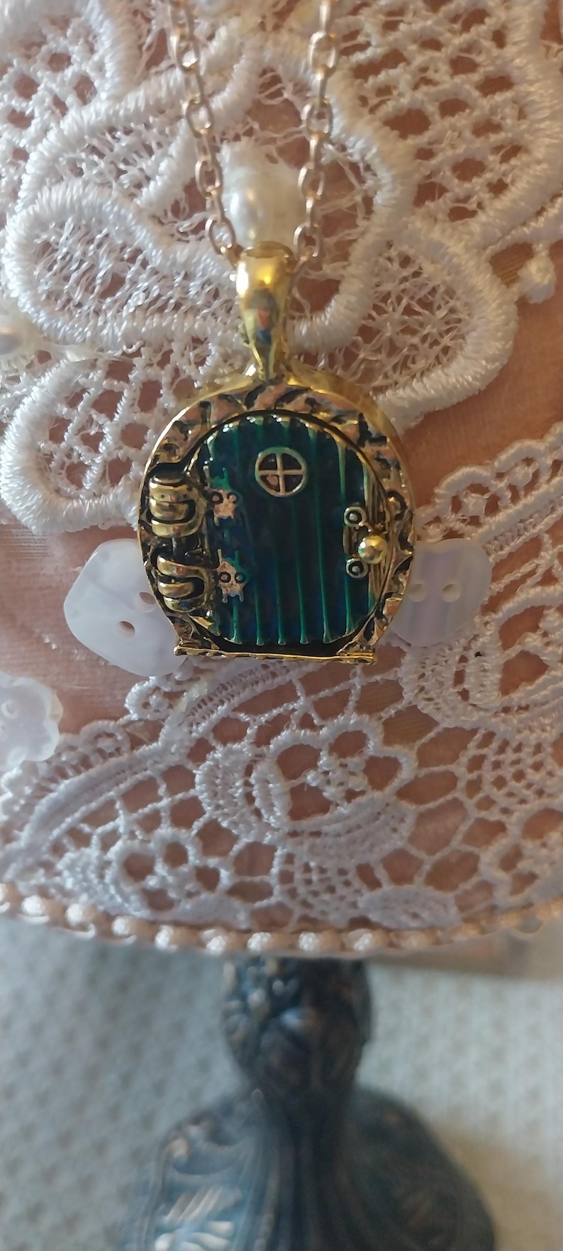 Pu&ograve; includere: Una collana con catena dorata con un ciondolo a forma di porta verde e dorata. La porta ha una piccola finestra rotonda e una maniglia dorata. La collana &egrave; su uno sfondo di pizzo bianco.