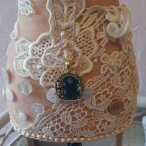 Pu&ograve; includere: Una collana con catena dorata con un ciondolo a forma di porta verde. La collana &egrave; esposta su un manichino rivestito di pizzo bianco. Il manichino &egrave; su un supporto nero con dettagli decorativi.