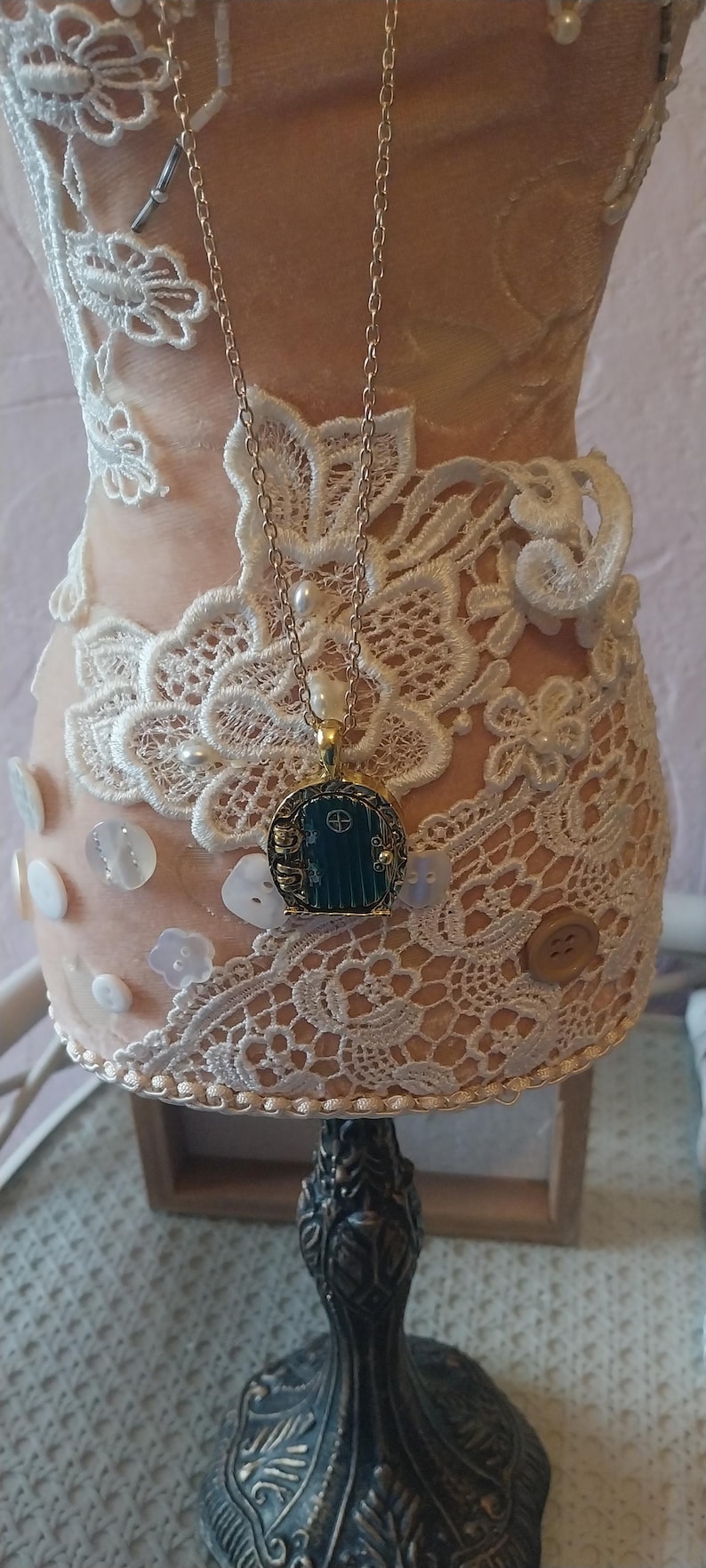 Pu&ograve; includere: Una collana con catena dorata con un ciondolo a forma di porta verde. La collana &egrave; esposta su un manichino rivestito di pizzo bianco. Il manichino &egrave; su un supporto nero con dettagli decorativi.