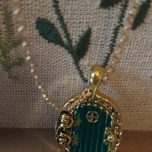 Pu&ograve; includere: Una collana con catena dorata con un ciondolo a forma di porta verde. La porta &egrave; circondata da una cornice dorata e ha una piccola finestra con un motivo a croce. Il ciondolo ha la forma di una porta di hobbit.