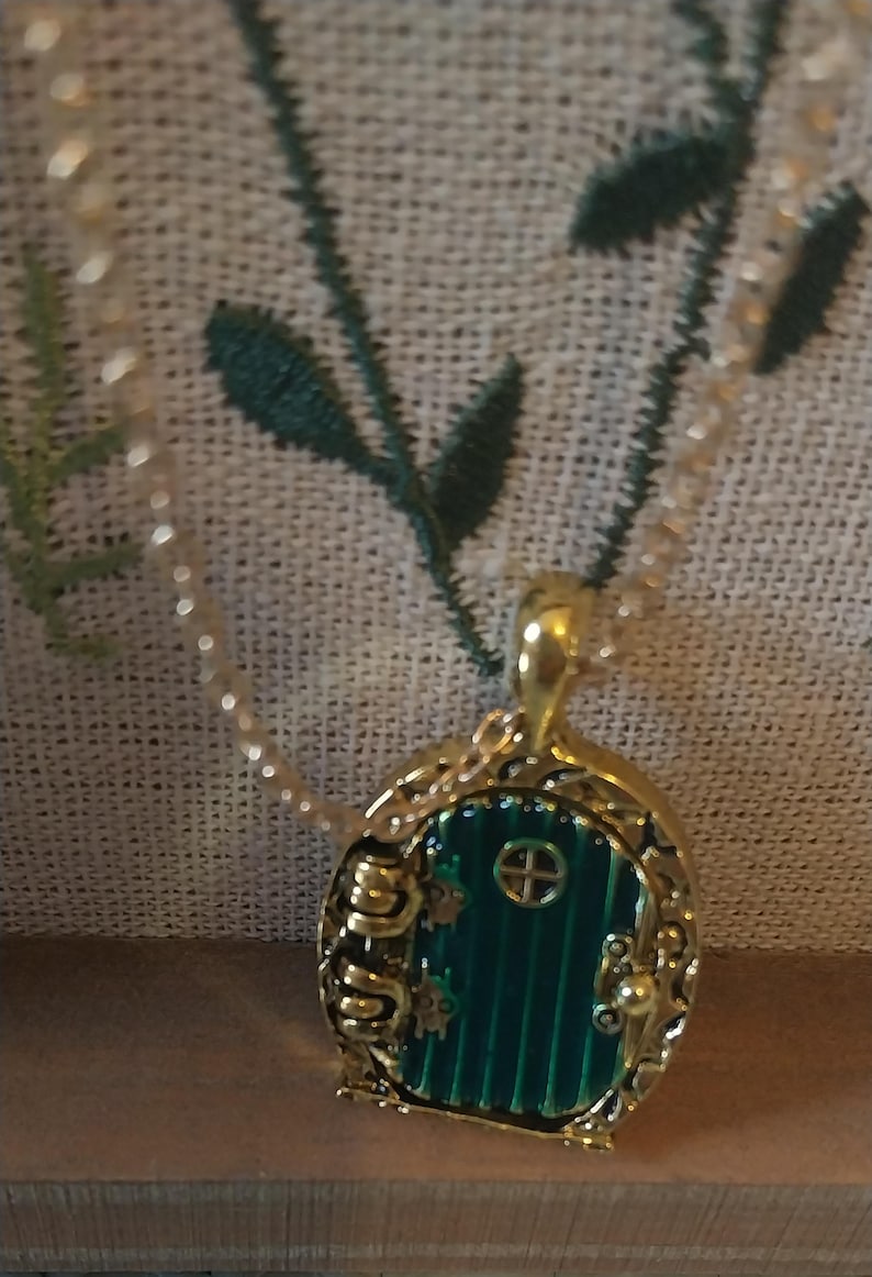 Pu&ograve; includere: Una collana con catena dorata con un ciondolo a forma di porta verde. La porta &egrave; circondata da una cornice dorata e ha una piccola finestra con un motivo a croce. Il ciondolo ha la forma di una porta di hobbit.