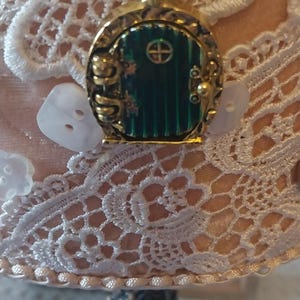 Pu&ograve; includere: Una collana dorata con un ciondolo a forma di porta verde. La porta ha una piccola finestra rotonda e una maniglia dorata. La collana &egrave; su uno sfondo di pizzo bianco.