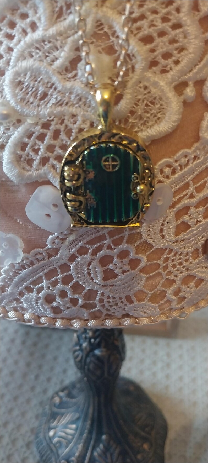 Pu&ograve; includere: Una collana dorata con un ciondolo a forma di porta verde. La porta ha una piccola finestra rotonda e una maniglia dorata. La collana &egrave; su uno sfondo di pizzo bianco.