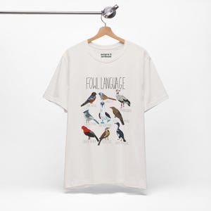 White unisex t-shirt with 'Fowl Language' bird design featuring tufted titmouse, blue-footed booby, and more. hanging from a wooden hanger, perfect for bird lovers and humor fans.