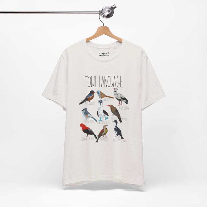 White unisex t-shirt with 'Fowl Language' bird design featuring tufted titmouse, blue-footed booby, and more. hanging from a wooden hanger, perfect for bird lovers and humor fans.