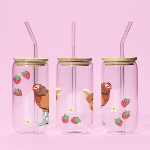 May include: Three clear glass tumblers with bamboo lids and glass straws. Each tumbler features a cartoon chicken wearing boots, strawberries, and flowers. The background is a solid pink.