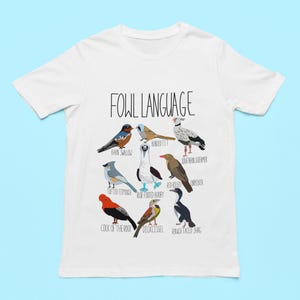 White unisex t-shirt with 'Fowl Language' bird design featuring tufted titmouse, blue-footed booby, and more. Flat lay on light blue background, perfect for bird lovers and humor fans.