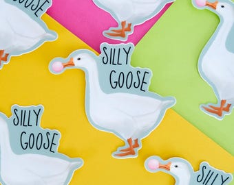 Puede incluir: Varias pegatinas troqueladas que muestran un ganso blanco con patas y pico de color naranja sosteniendo una burbuja de chicle rosa. Cada pegatina incluye el texto negro SILLY GOOSE impreso en una fuente informal sobre un fondo azul claro.