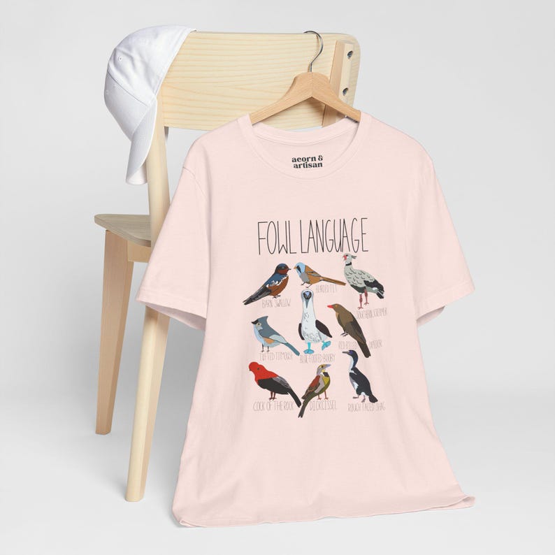 Soft Pink unisex t-shirt with 'Fowl Language' bird design featuring tufted titmouse, blue-footed booby, and more. Shirt hanging on wooden chair, perfect for bird lovers and humor fans.