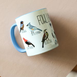 May include: White ceramic mug with a blue rim and handle. The mug features a colorful illustration of different birds with their names written below them. The text "Fowl Language" is printed in black at the top of the mug.