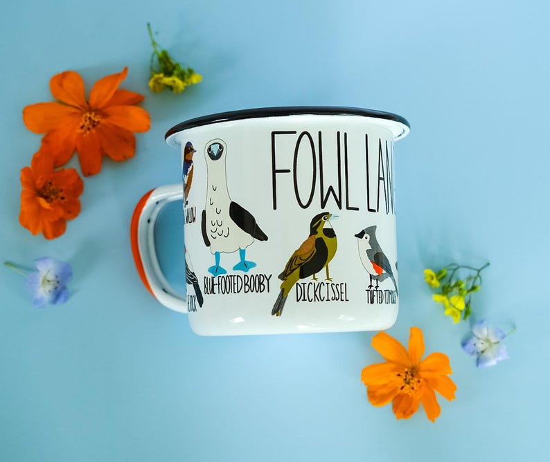 May include: White enamel mug with a black rim and handle, featuring illustrations of various birds and the text "FOWL LAND". The mug has an orange accent on the handle and is surrounded by orange flowers and blue flowers.