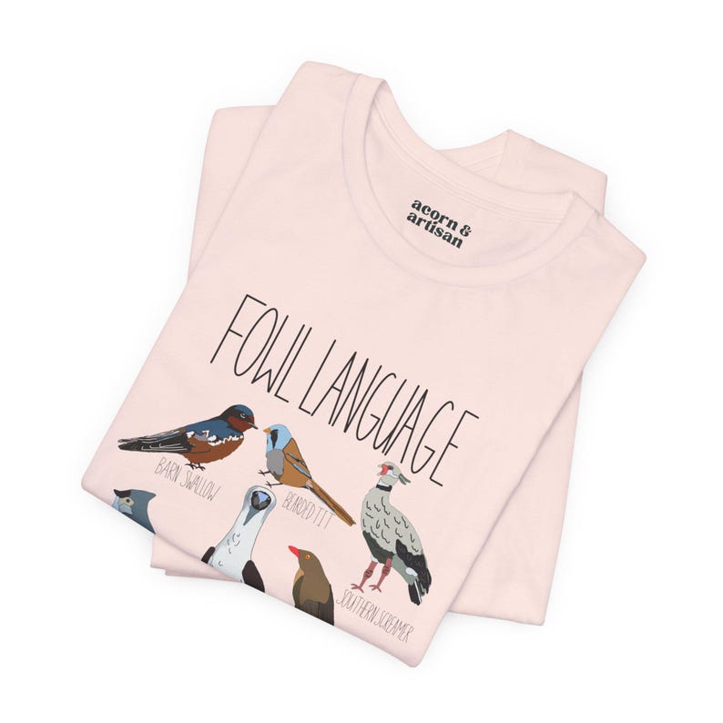 Soft Pink unisex t-shirt with 'Fowl Language' bird design featuring tufted titmouse, blue-footed booby, and more. Shirt folded in pile to show close up detail and tagless design.