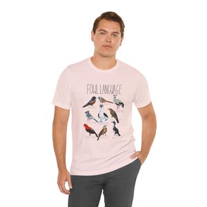 Soft pink unisex t-shirt with 'Fowl Language' bird design featuring tufted titmouse, blue-footed booby, and more on male model.