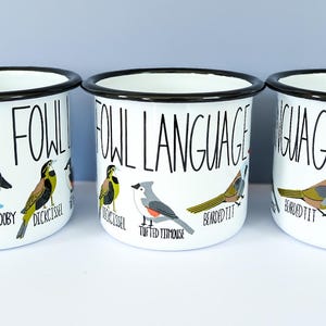 May include: Three white enamel mugs with black rims and orange handles. Each mug features the text "FOWL LANGUAGE" and illustrations of various birds. The mugs are designed for beverages.