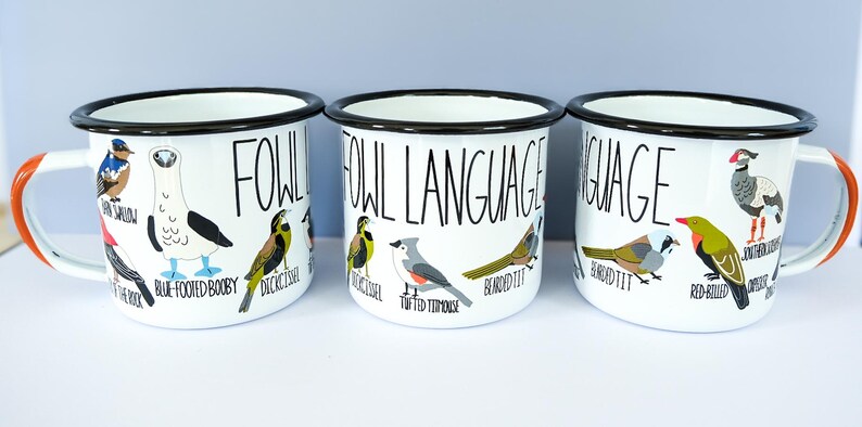 May include: Three white enamel mugs with black rims and orange handles. Each mug features the text "FOWL LANGUAGE" and illustrations of various birds. The mugs are designed for beverages.