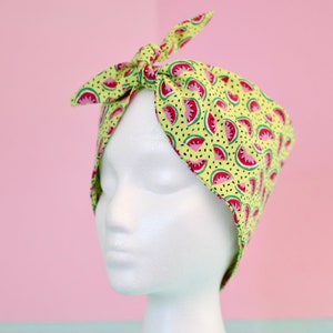 May include: A green headband with a watermelon print. The headband is tied in a knot at the top and is worn on a white mannequin head.