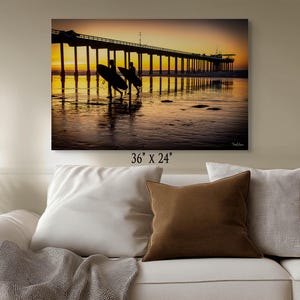 May include: A canvas print of a pier at sunset. Two surfers walk towards the water, carrying their surfboards. The sky is a gradient of orange and yellow, reflecting on the wet sand. The print measures 36" x 24".