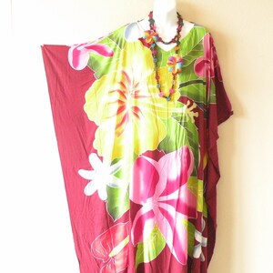 May include: A loose-fitting kaftan dress in deep burgundy featuring a vibrant floral print with large yellow hibiscus and pink plumeria flowers, green leaves, and white accents. The garment is paired with a long beaded necklace with flower charms.
