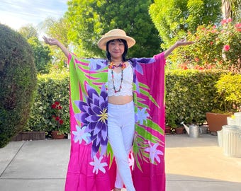 Pode incluir: Um kimono vibrante em tons de rosa, verde e roxo, adornado com padrões florais. É usado com um top e calças brancas, um chapéu de palha e um colar. Um traje de verão.