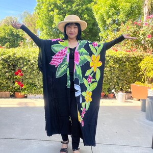 May include: A black kimono-style robe with a vibrant floral design in pink, yellow, and green. The robe is worn with a straw hat and black clothing. The background features lush greenery and a sunny sky.