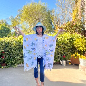 Op de afbeelding: Witte kimono met een tropische print met schildpadden, bladeren en fruit. De kimono heeft een kwastjesrand en wordt gedragen met een witte top, een blauwe spijkerbroek en een lichtblauwe hoed. De achtergrond omvat groen en een zonnige hemel.