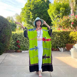 May include: A lime green kimono with black trim and a fish design. The kimono is worn with a white top and pants, a straw hat, and a colorful necklace. The background features lush greenery and a sunny sky.